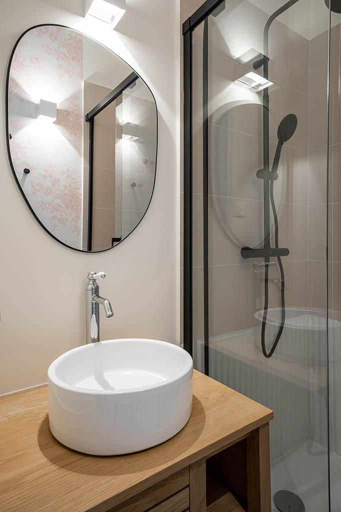 Espace salle d'eau dans chambre d'amis, douche paroi vitrée et robinetterie noire, meuble vasque bois et vasque blanche, miroir noire forme organique