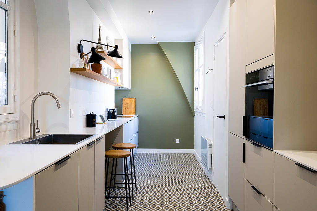 Linéaire cuisine épurée façades beige, plan de travail Corian blanc, étagères bois sur mesure, poignées modernes noires, mur du fond vert, sol carrelage graphique